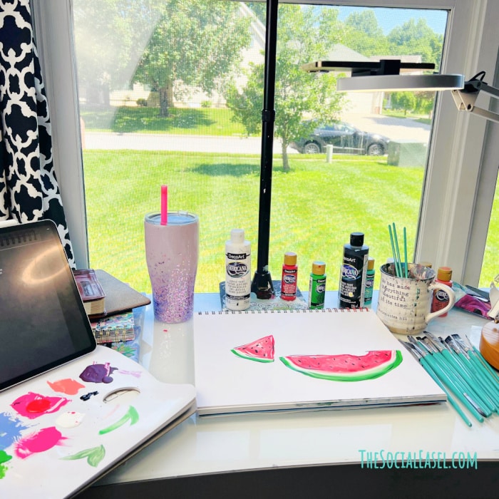Christie's desk with painting supplies, laptop, and watermelon slice painting. Window to the outdoors