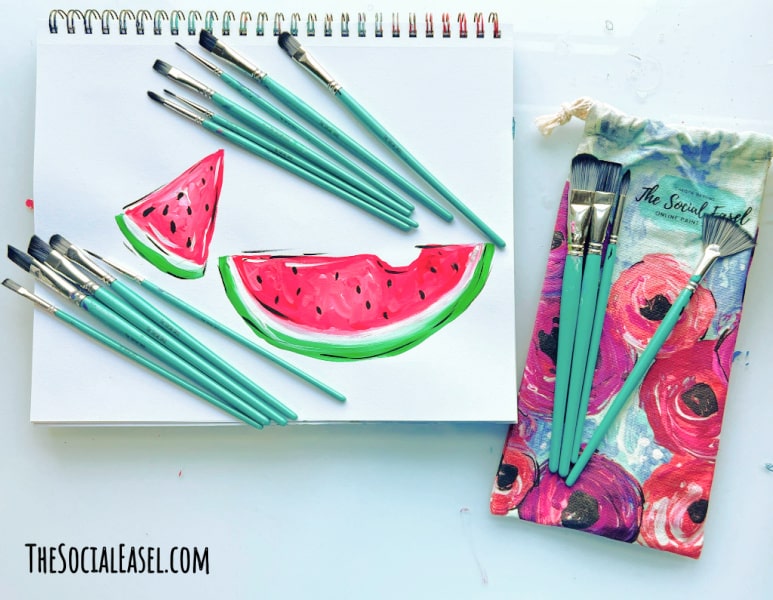 Watermelon slices painted on a paper pad surrounded by aqua paint brushes with a floral bag