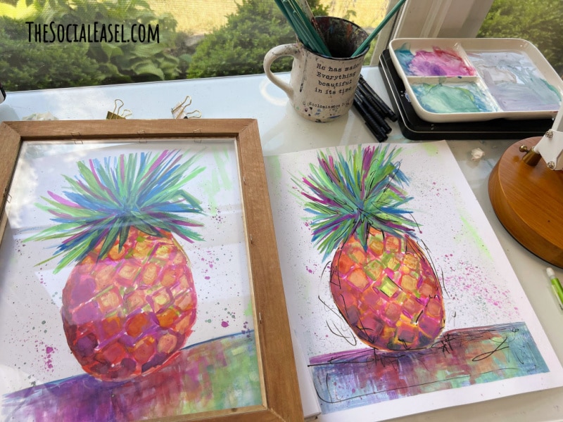 two watercolor pineapple paintings on the art desk one set inside a frame