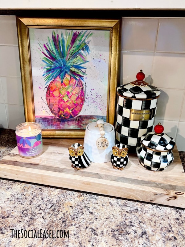 Funky watercolor pineapple painting in a gold frame staged on my kitchen counter with black and white check jars