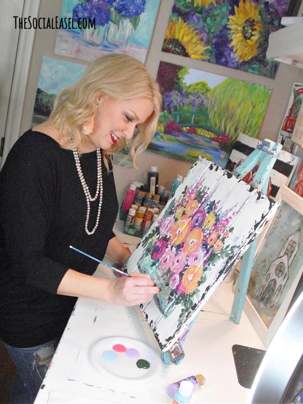 Christie Hawkins painting country flowers on a wood pallet board in her studio surrounded by her artwork