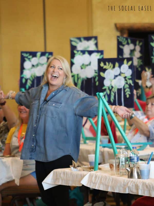 Christie Hawkins with arms out happily looking at all the progress of Bold Bloom paintings at her live event.