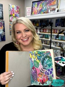 Christie Hawkins holding her art journal. Showing an abstract art design with yellow, green, purple, orange.