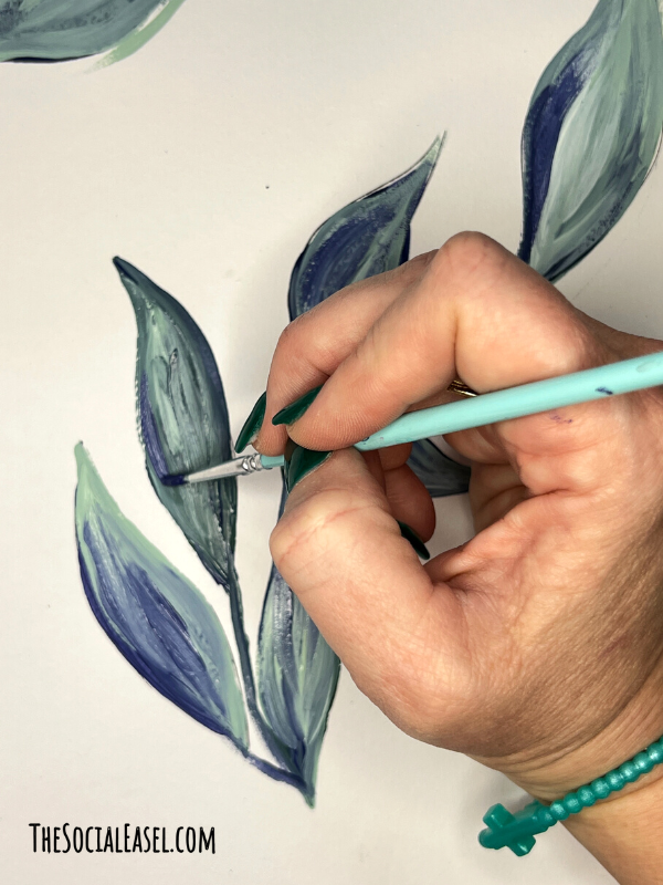 Christie holding a small paintbrush adding darker detail to the outer edge of the middle leaf.