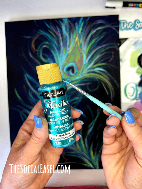 A bottle of Deco Art Metallics paint in Peacock pearl with a paint brush with some paint on it. A painted colorful peacock feather on canvas in the back ground. 