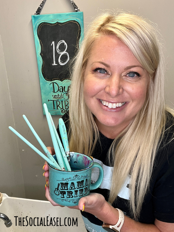 Christie Hawkins holding a mug she uses to rinse her paintbrushes. 6 paintbrushes with teal handles.