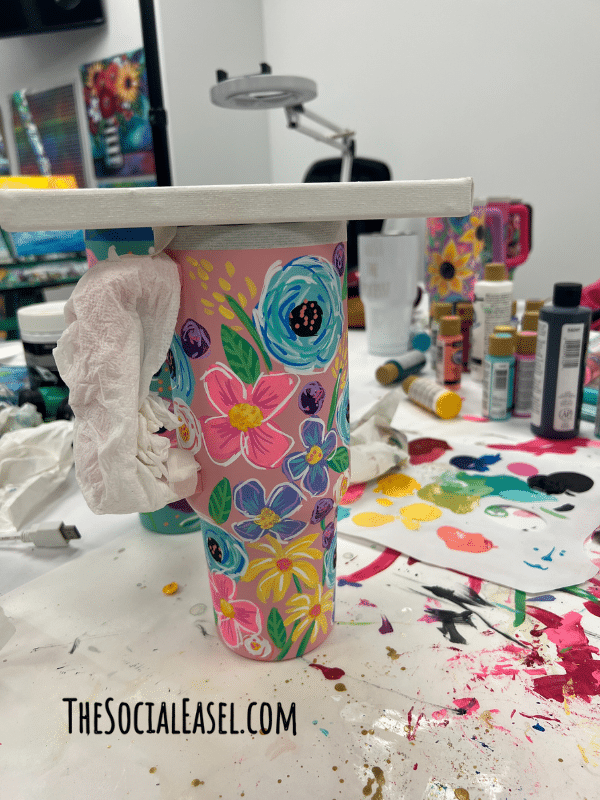 A Stanley Style Cup (pink) with hand painted colorful blue, pink, purple flowers. The handle, and top edge are wrapped, and the top is covered to protect it when putting on a sealer. 