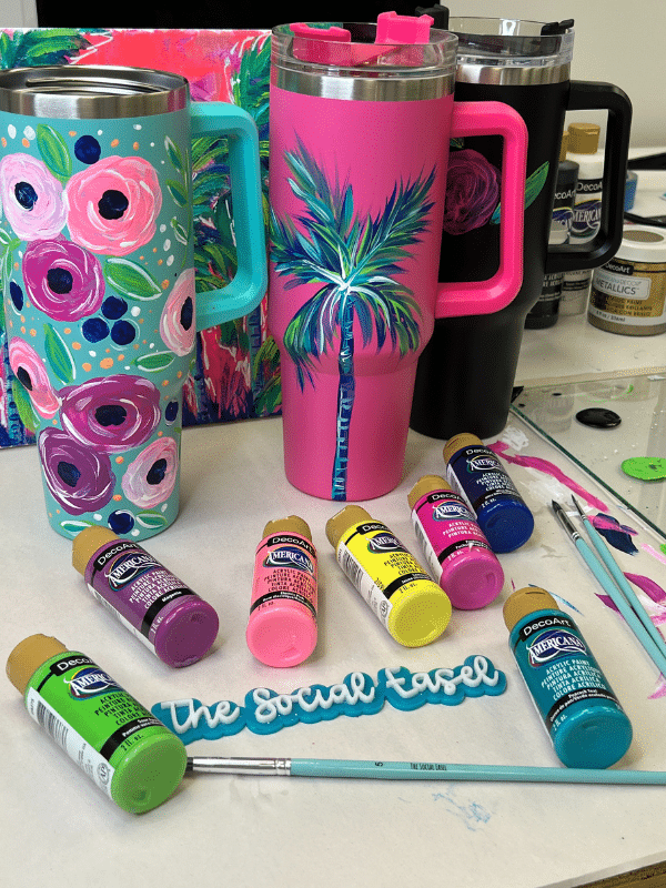 3 Stanley style drinking cups hand painted. The first one is teal with bold blooms of light and dark pink, The Second is pink and has a colorful palm tree with blue, green, purple and white. And the third one is a black cup with flowers. 