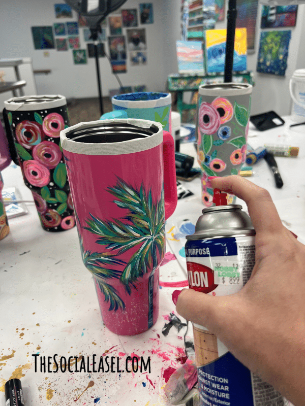 Hot pink cup with a colorful palm tree. Christie Hawkins holding a spray sealer to protect the hand painted cup. 