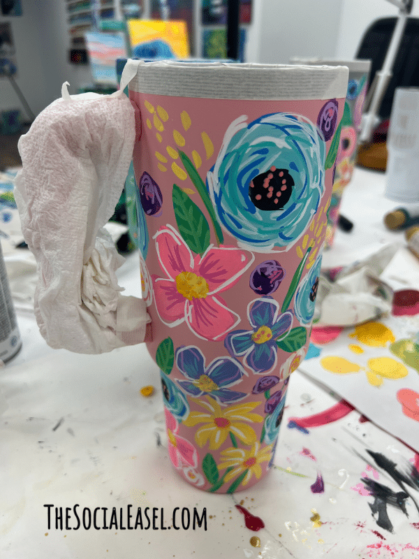 A Hand painted cup with colorful blooms. The handle is covered in paper towel to protect it when using a spray sealer. 