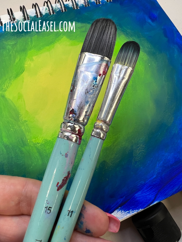 Small and large Filbert brushes out of Christie's signature paint brush set. The background in an ombre design on a mixed media pad. Blue, green, and yellow.