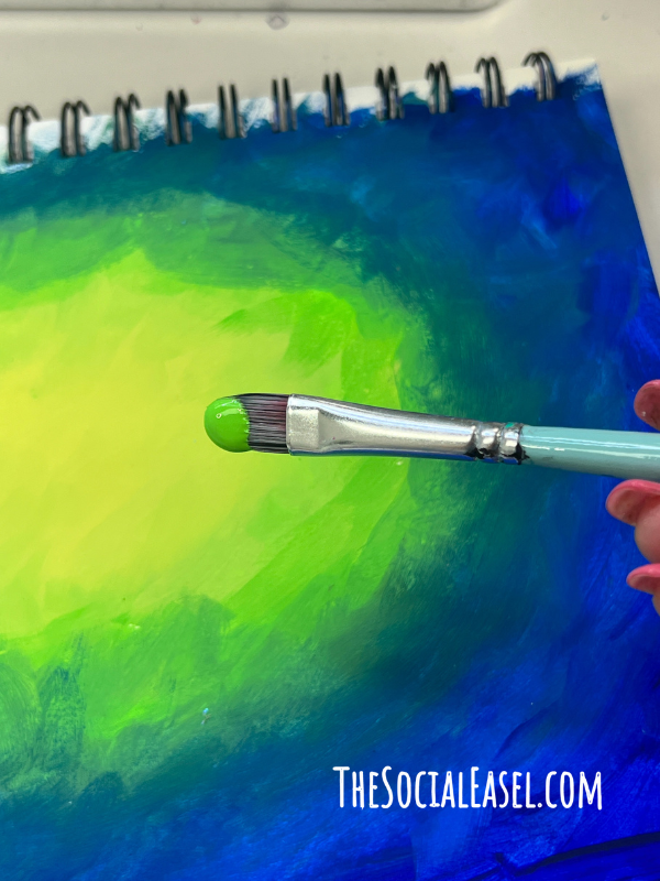 A Paint brush with bright green acrylic paint on it. Back ground is a mixed media pad with bright blue edges, green, yellow in the center. Blending edges connecting the colors. 