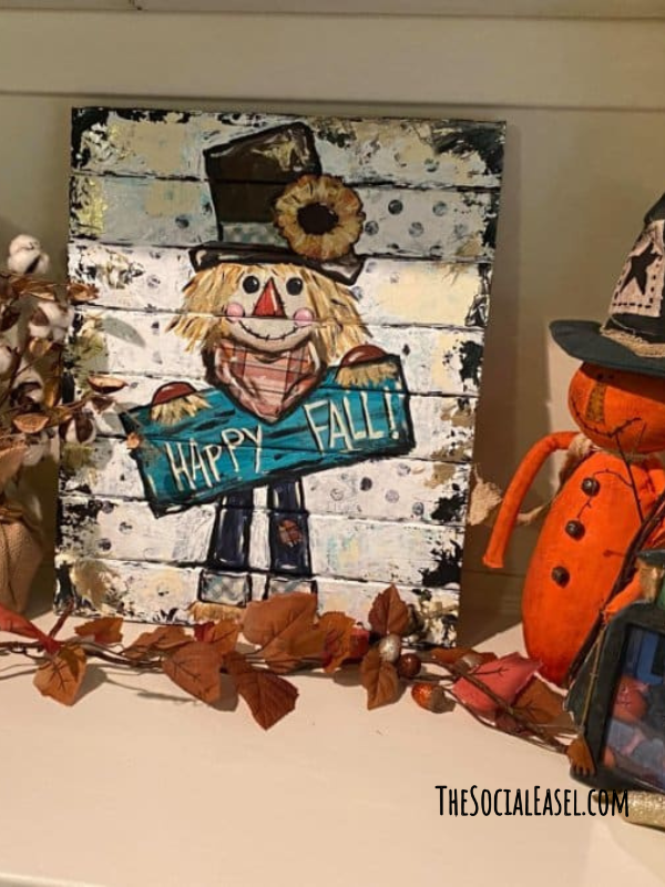 A scarecrow painting on a wood palette with a Happy Fall sign surrounded by fall decor. 