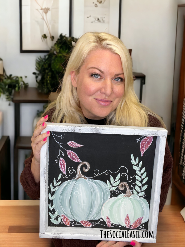 Christie Hawkins holding a wooden whitewashed frame with a painting of two pumpkins. The background is black, and the pumpkins are a gray blue, and light soft green. There are some vines and leaves surrounding the pumpkins. 