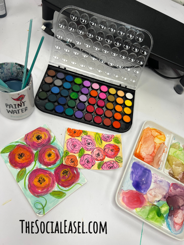 A display of watercolors, paintbrushes in a mug of water, 2 watercolor painted postcards, and a paint tray.